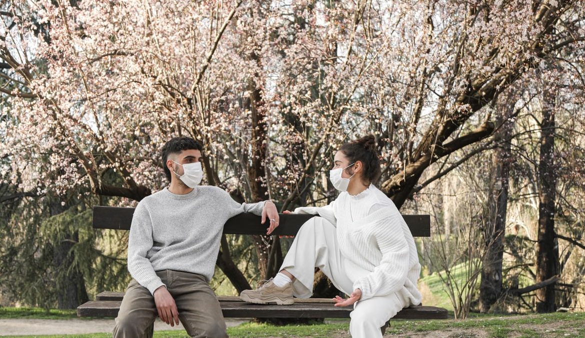 Young couple wearing face masks sitting on a park bench, practicing social distancing during the pandemic.